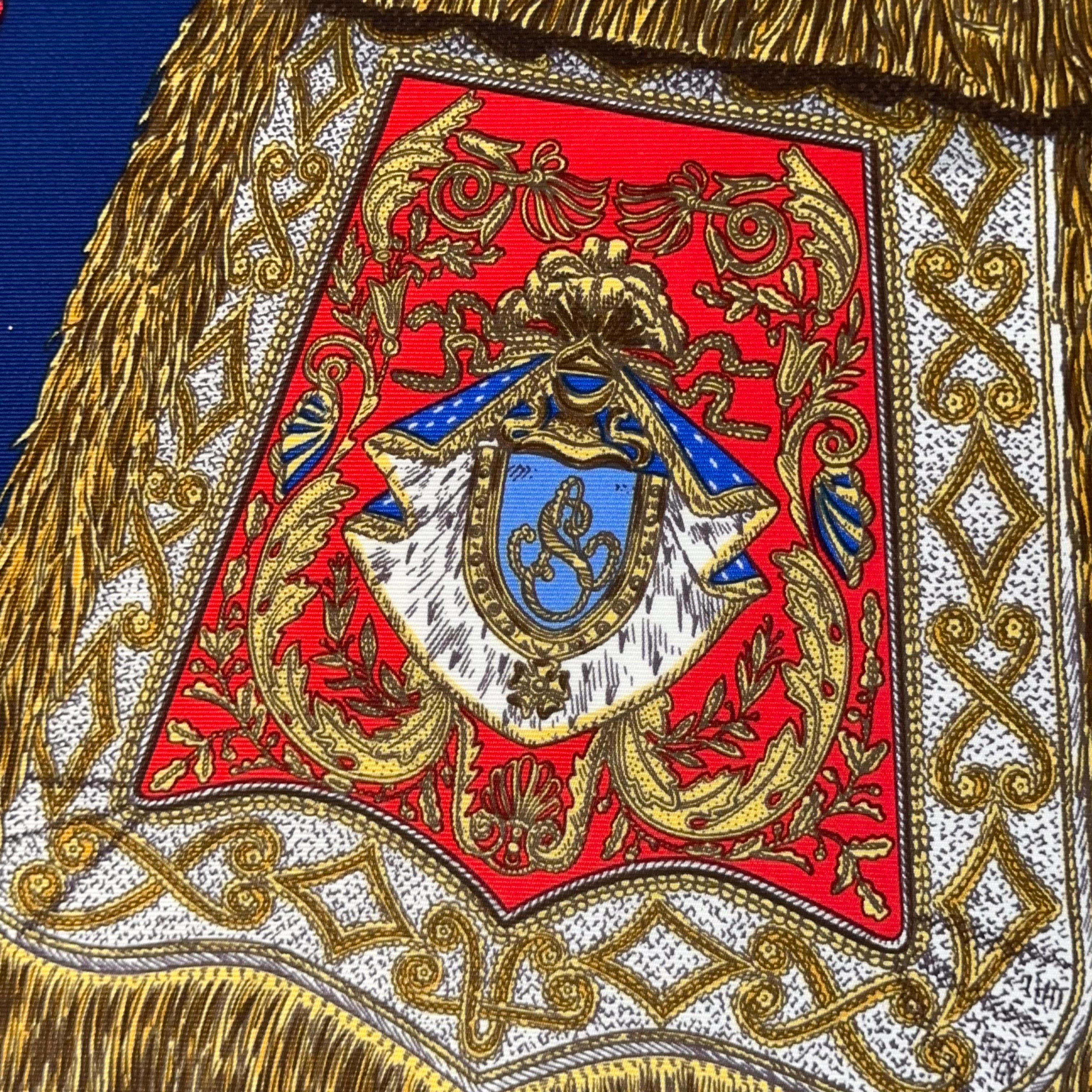 Gros plan sur une sabretache latérale rouge et or, ornée d'un monogramme bleu, montrant la richesse des passementeries dessinées par Metz.