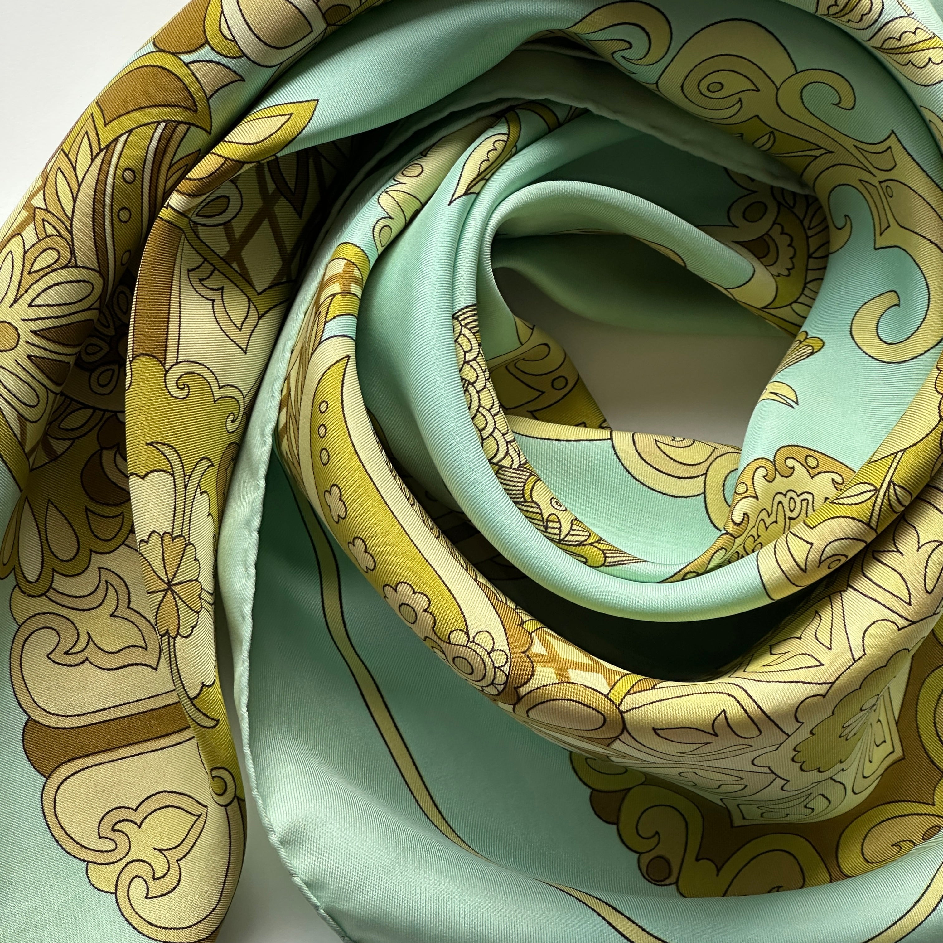 Le carré Cendrillon drapé en tourbillon, mettant en valeur l'harmonie des couleurs vert, or et olive pour un look vintage.